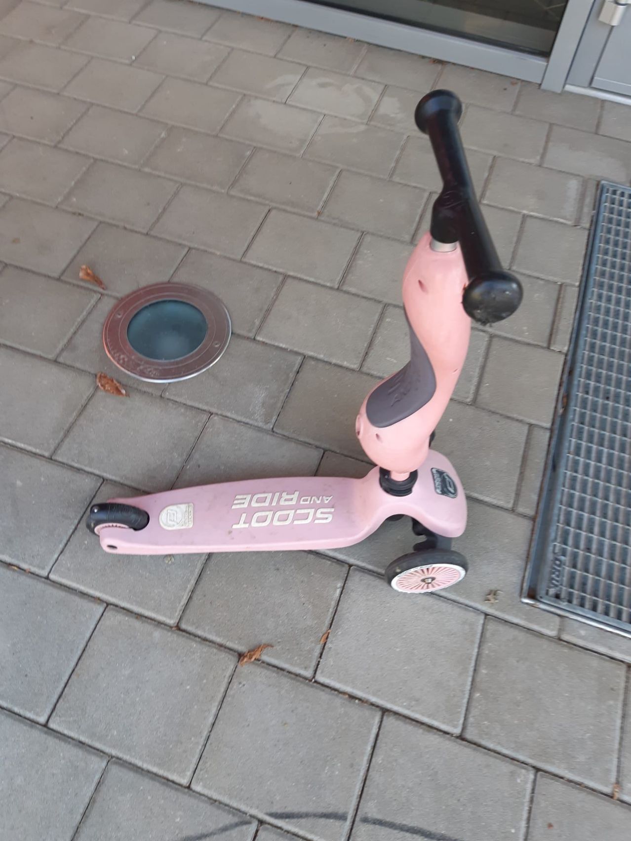 Ein rosafarbener Roller mit der Marke Scoot liegt auf einem Ziegelboden. Ein runder Abflussdeckel befindet sich in der Nähe.