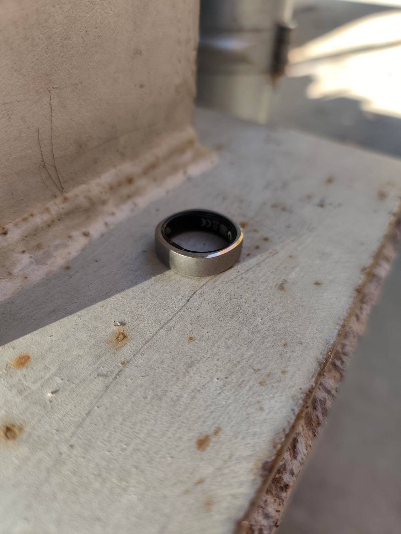 Ein metallischer Ring mit schwarzer und silberner Färbung liegt auf einer Betonoberfläche mit einigen Rostflecken.