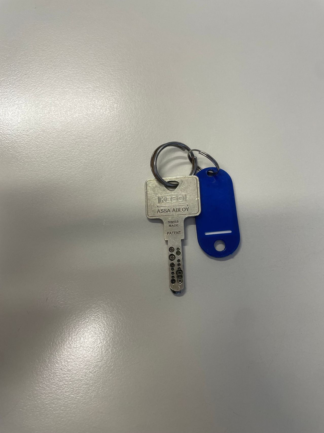 A silver key and a blue tag are displayed on a white surface. The key has the word ASSA ABLOY on it.