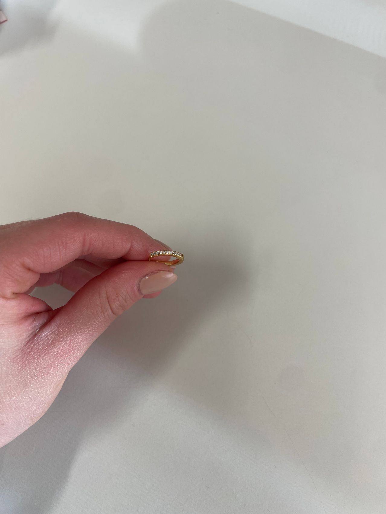 Eine Hand hält einen goldenen Ring mit kleinen Diamanten. Der Hintergrund ist eine schlichte weiße Oberfläche.