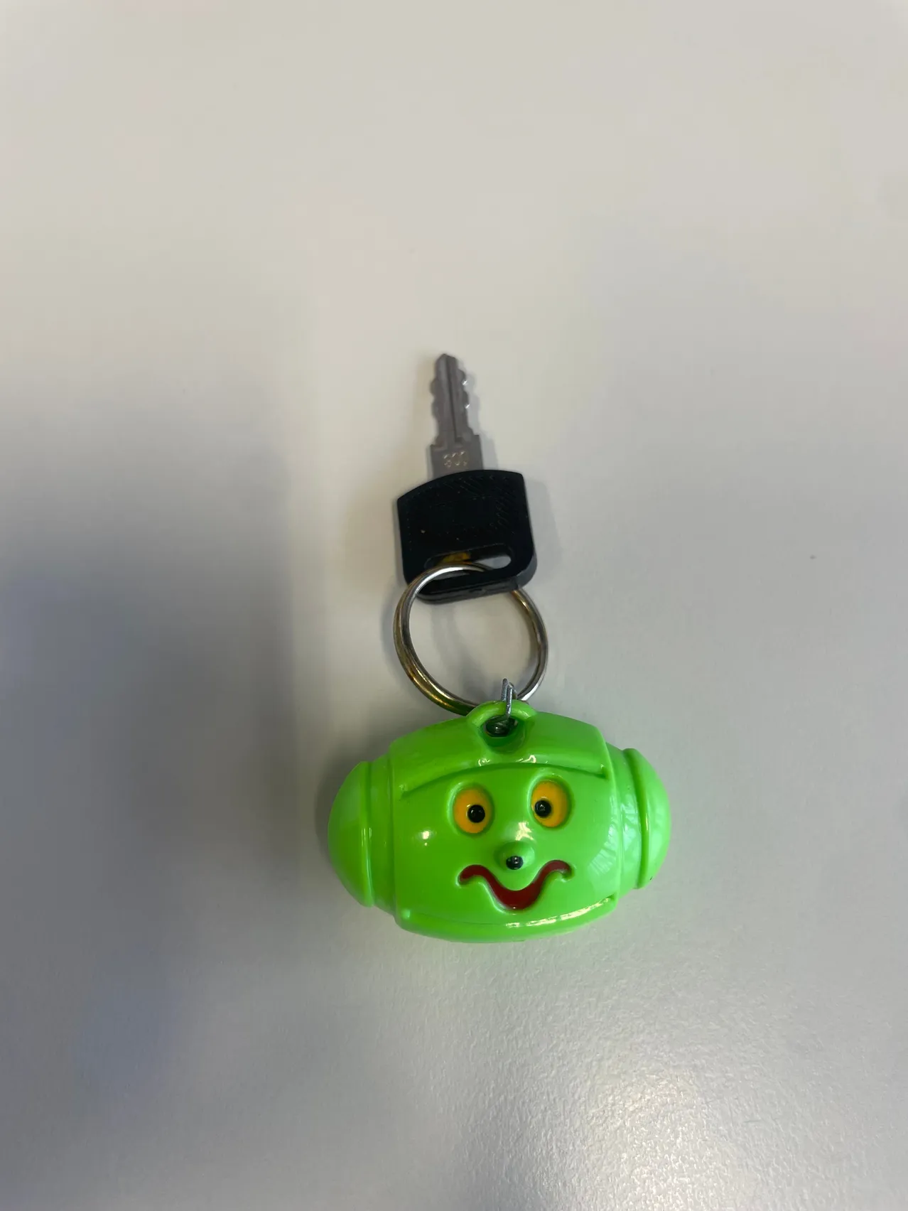 Ein grüner Schlüsselanhänger in Form eines Roboters mit gelben Augen und einem roten Mund. Er hat einen Schlüssel, der an einem silbernen Ring befestigt ist.