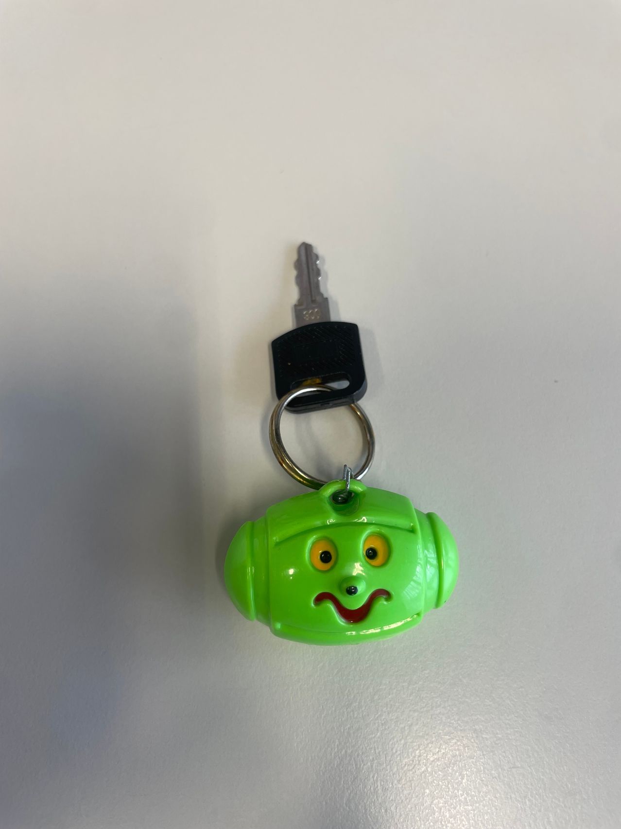 A green keychain shaped like a robot with yellow eyes and a red mouth. It has a key attached to a silver ring.