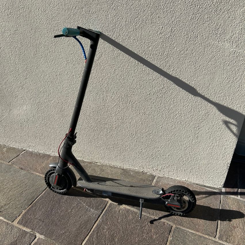Ein schwarzer Elektroroller steht auf einem steinernen Bürgersteig neben einer strukturierten weißen Wand und wirft einen Schatten.