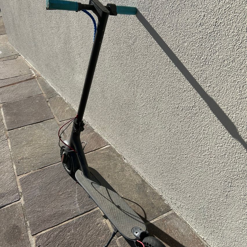 Ein schwarzer Elektroroller mit türkisfarbenen Griffen steht auf einem Steinpfad neben einer Wand.