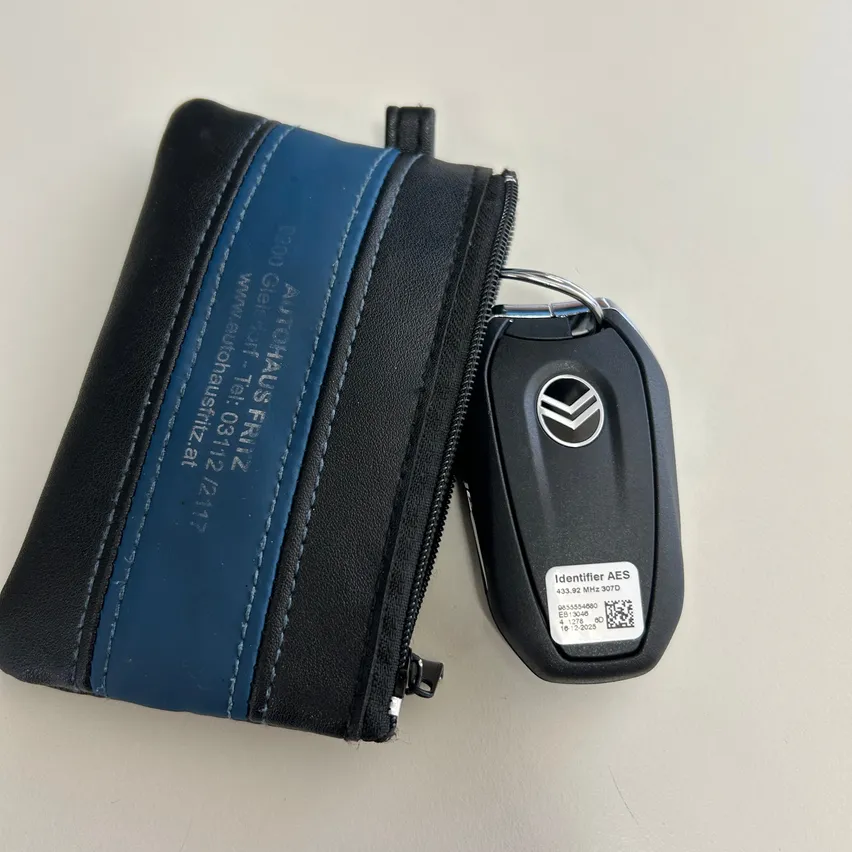 Ein schwarz-blauer Lederschlüsselanhänger mit einem Volkswagen-Schlüsselanhänger. Der Schlüsselanhänger hat einen Reißverschluss und einen blauen Streifen mit weißem Text. Der Schlüsselanhänger hat ein schwarzes und silbernes Design mit einem QR-Code und dem Text 'Identifier AES'.