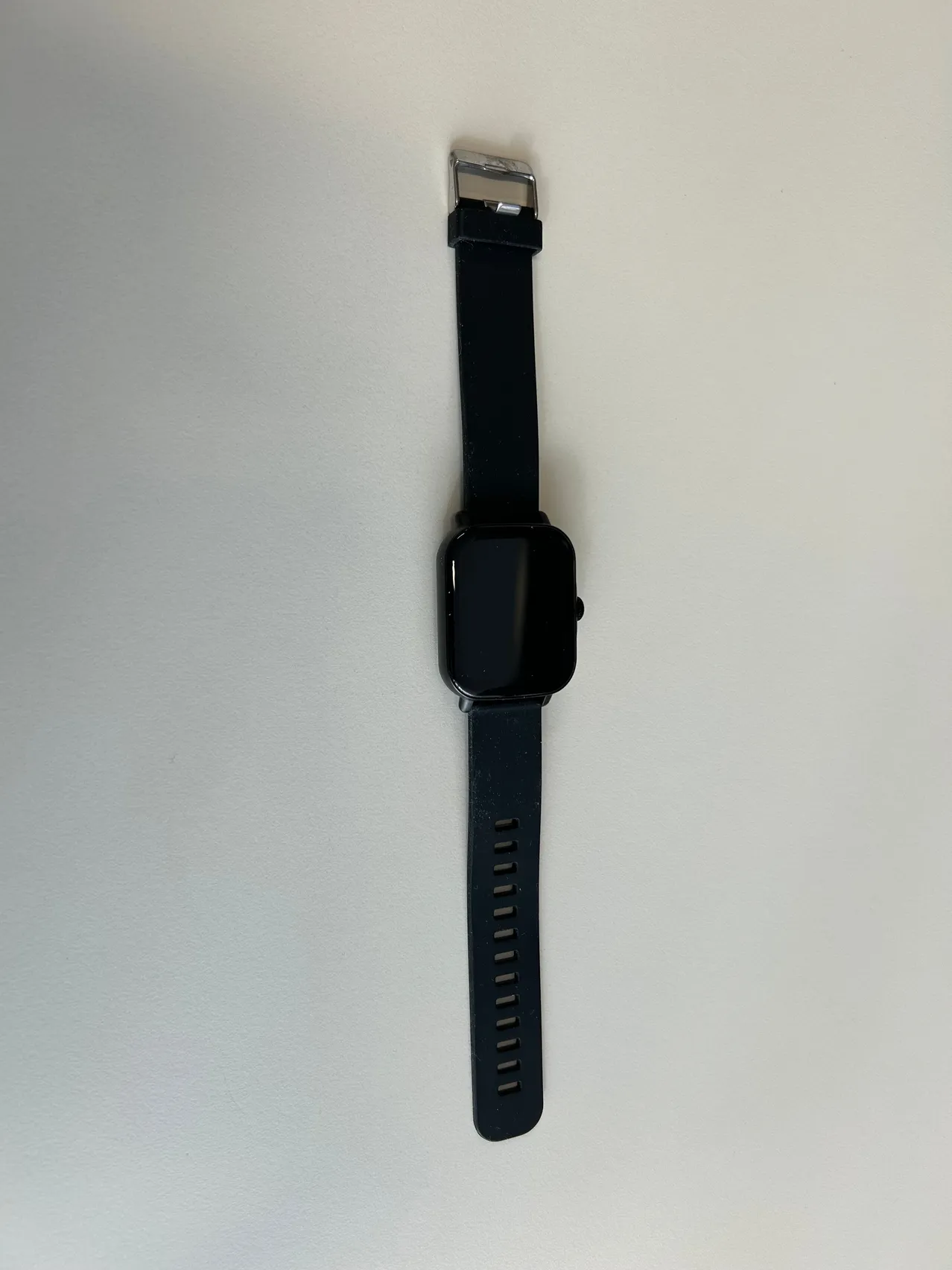 Eine schwarze Smartwatch mit schwarzem Armband ist an einer weißen Wand montiert. Das Uhrengesicht ist ausgeschaltet.