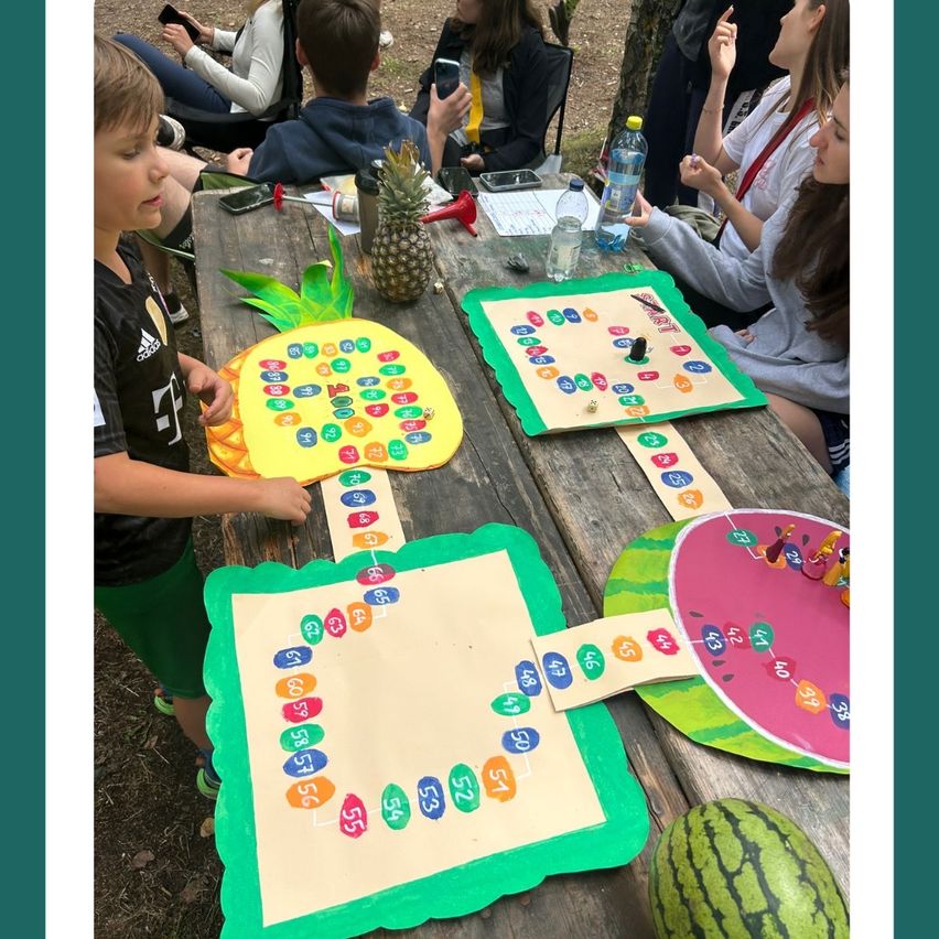 Eine Gruppe von Kindern und Erwachsenen spielt ein Brettspiel auf einem Picknicktisch mit Fruchtdekorationen.
