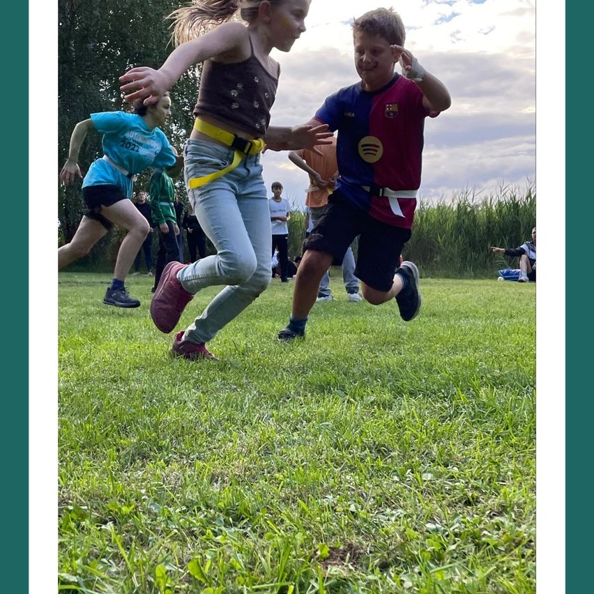 Mehrere Kinder rennen auf einem Rasenfeld. Ein Mädchen vorne mit einem gelben Gürtel wird von einem Jungen in einem blau-roten Shirt verfolgt. Dahinter stehen andere Kinder und beobachten.