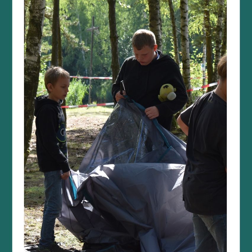 Drei Jungen bauen ein Zelt in einem Wald auf. Der Junge in der Mitte justiert das Zelt. Der Junge auf der linken Seite beobachtet. Der Junge auf der rechten Seite schaut etwas an.