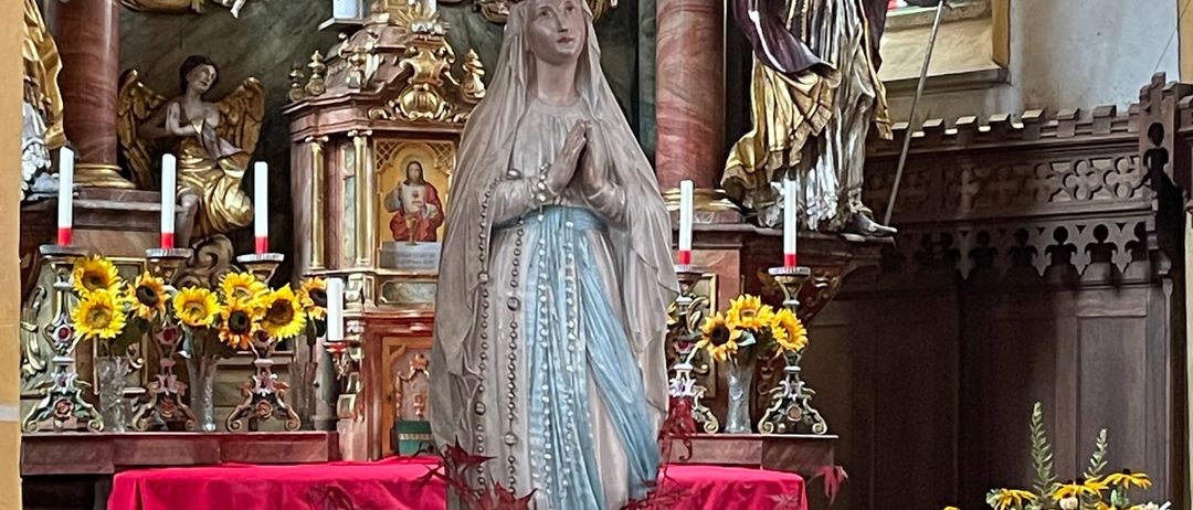 Ein Altar mit einer Statue der Jungfrau Maria und eines Bischofs, geschmückt mit Blumen, Kerzen und dekorativen Elementen, steht in einer Kirche.