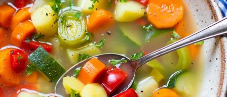 Eine Schüssel mit Gemüsesuppe gefüllt mit gewürfelten Karotten, Tomaten, Gurken und anderen Gemüsesorten. Ein Löffel wird benutzt, um die Suppe zu schöpfen.