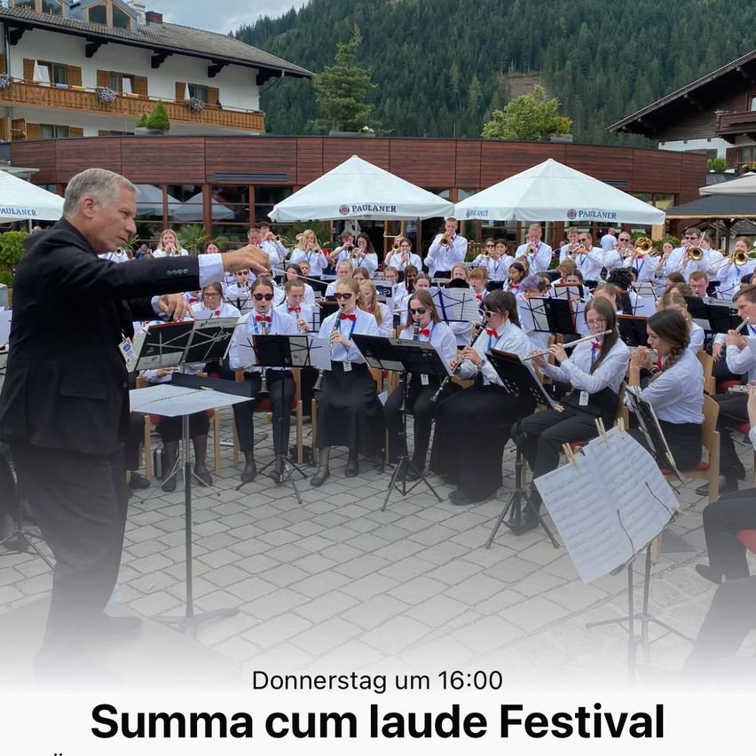 Ein Dirigent führt eine Brass Band im Freien auf einem gepflasterten Boden. Dahinter ist ein Gebäude sichtbar, mit Bergen im Hintergrund. Textüberlagerung: Donnerstag um 16:00 Uhr und Summa cum laude Festival.