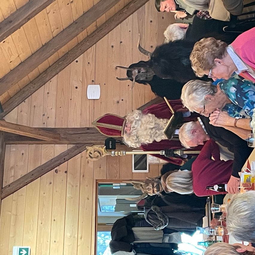 A group of people gather in a room with wooden walls. Some are wearing glasses and hats. One man is seated and looking at a woman. There is a black statue with horns in the room.