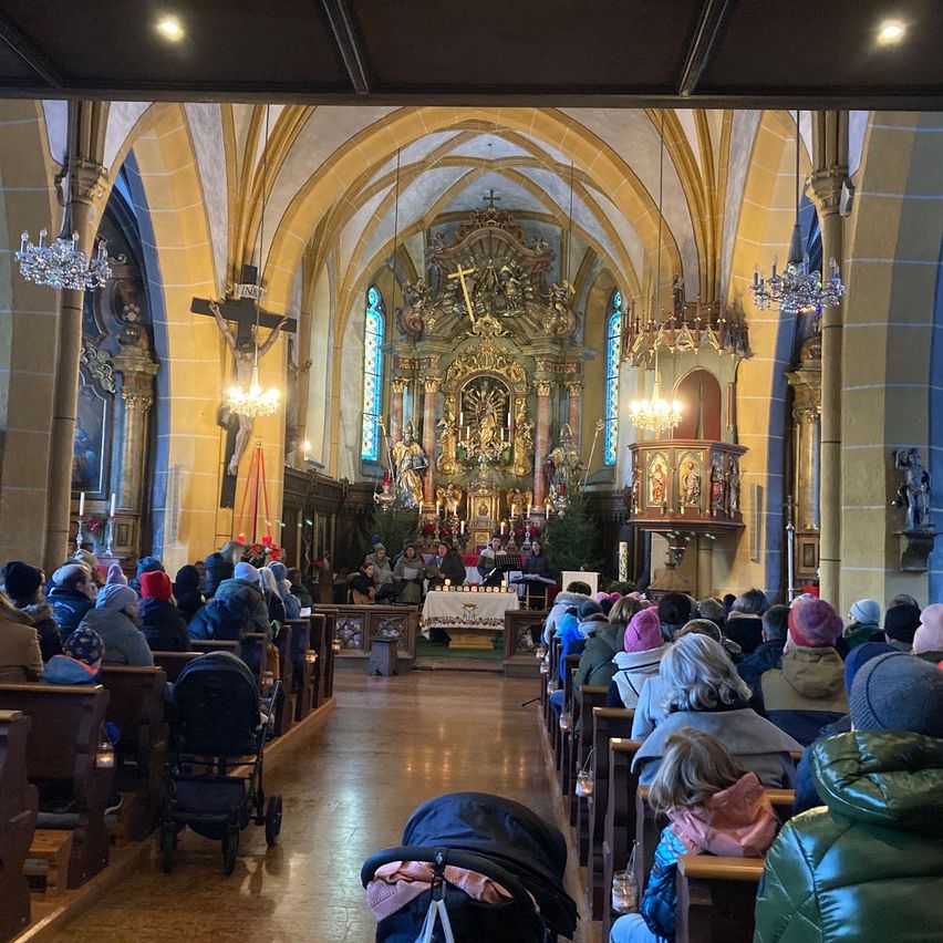 Im Inneren einer Kirche sitzen viele Menschen auf Kirchenbänken. Der Altar ist mit Kerzen, einem Tisch und einem Kreuz geschmückt. Die Decke ist mit Kronleuchtern und Säulen verziert. Ein Kinderwagen steht vorne auf dem Boden.