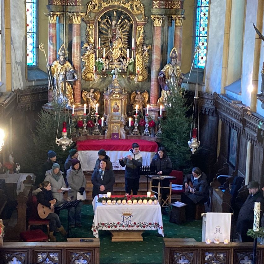 Eine Gruppe von Menschen tritt in einer Kirche auf, mit Weihnachtsbäumen, Kerzen und Musikinstrumenten im Vordergrund. Dahinter befindet sich ein reich verzierter Altar mit Statuen und aufwendigen Dekorationen.