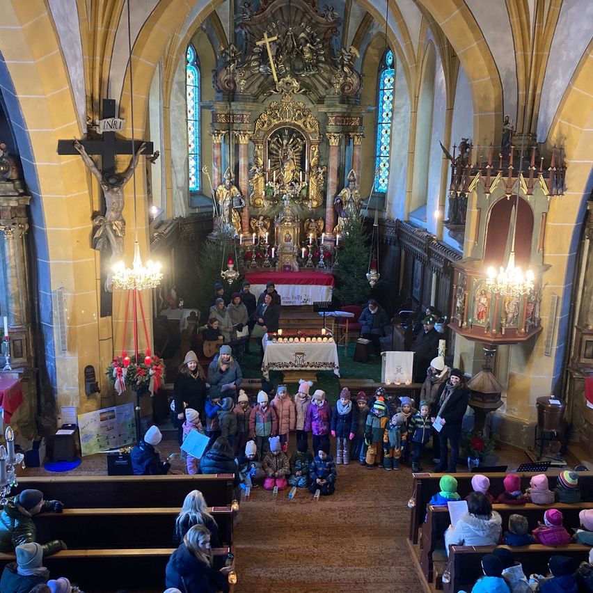 Eine Gruppe von Menschen versammelt sich in einer Kirche, einschließlich Kinder in Winterkleidung, für eine festliche Veranstaltung. Der Altar und die Umgebung sind mit Kerzen und Weihnachtsdekoration geschmückt.
