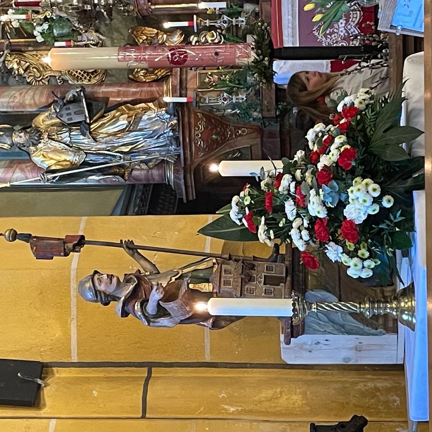 A church interior with statues and flowers. One statue holds a sword and a candle. Another statue with a shield is on the wall. Candles and flowers are placed in the foreground.