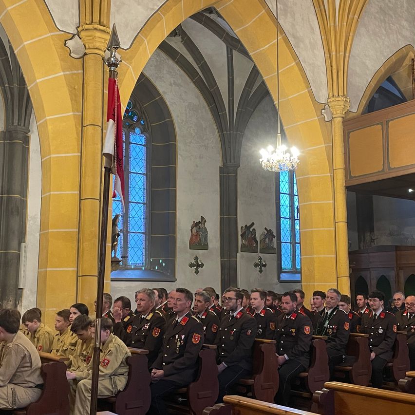 A military ceremony inside a church with soldiers in uniform sitting on benches under a flag. The walls are yellow, and stained glass windows illuminate the room.