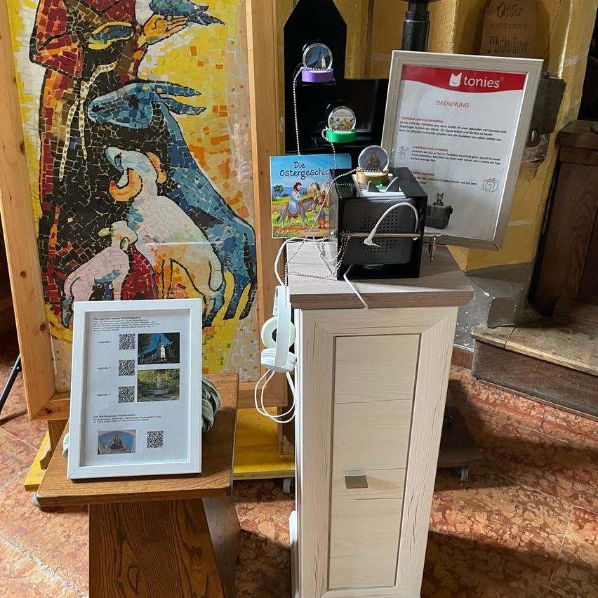 Ein kleiner Holztisch trägt ein gerahmtes Bild mit QR-Codes. Daneben zeigt ein weißer Schrank ein kleines elektronisches Gerät mit verschiedenen Gegenständen darauf. Dahinter zeigt ein farbenfrohes Mosaikbild eine Szene mit einer Person und Tieren. Ein rotes Schild mit dem Wort 'tonies' ist an der Wand befestigt.