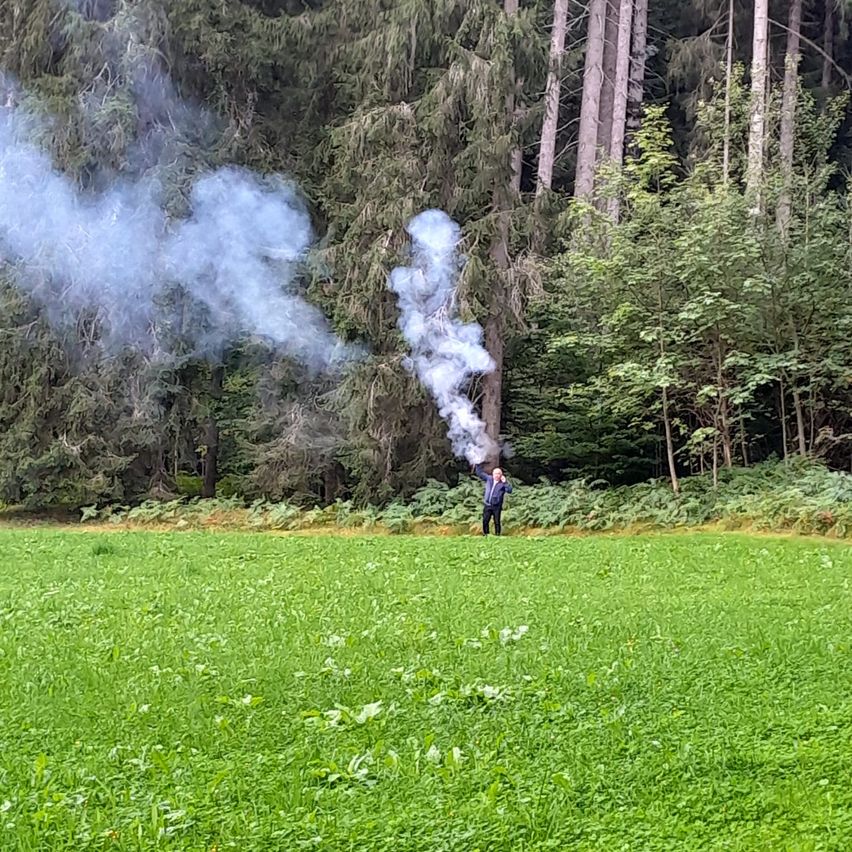 Eine Person steht auf einer Wiese mit Bäumen im Hintergrund und lässt Rauch aus einem Gerät entweichen.