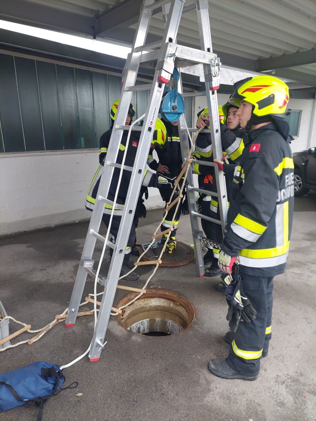 Mehrere Feuerwehrleute in Uniform klettern auf Leitern in der Nähe eines großen offenen Schachtes.