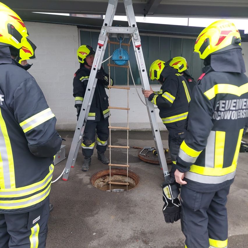 Feuerwehrleute in gelben und schwarzen Uniformen mit Helmen stehen um eine Leiter über einem offenen Kanaldeckel, einer klettert hinab.