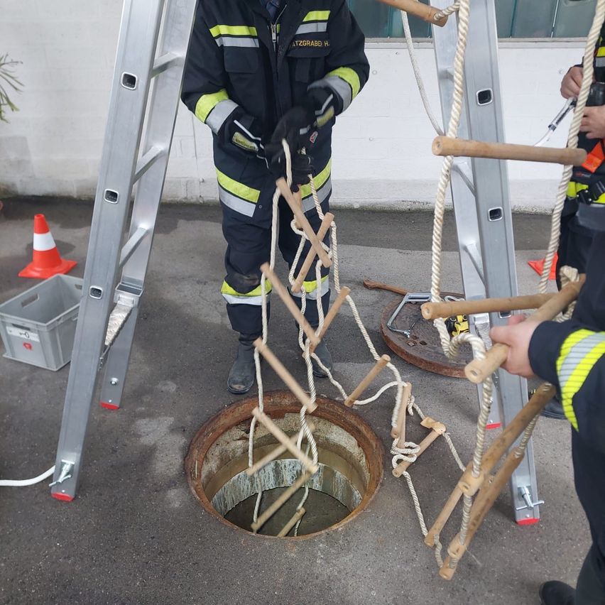 Zwei Feuerwehrleute senken eine Bambusleiter in einen Schacht und befestigen sie mit Seilen. Ein Feuerwehrmann trägt Handschuhe und beide tragen reflektierende Westen.