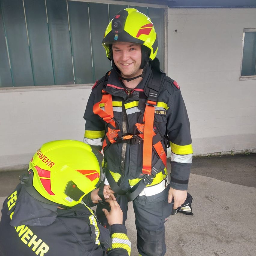 Zwei Feuerwehrleute in gelben Helmen, einer hält den Gürtel des anderen, stehen vor einem Gebäude mit Fenstern und einer Tür.