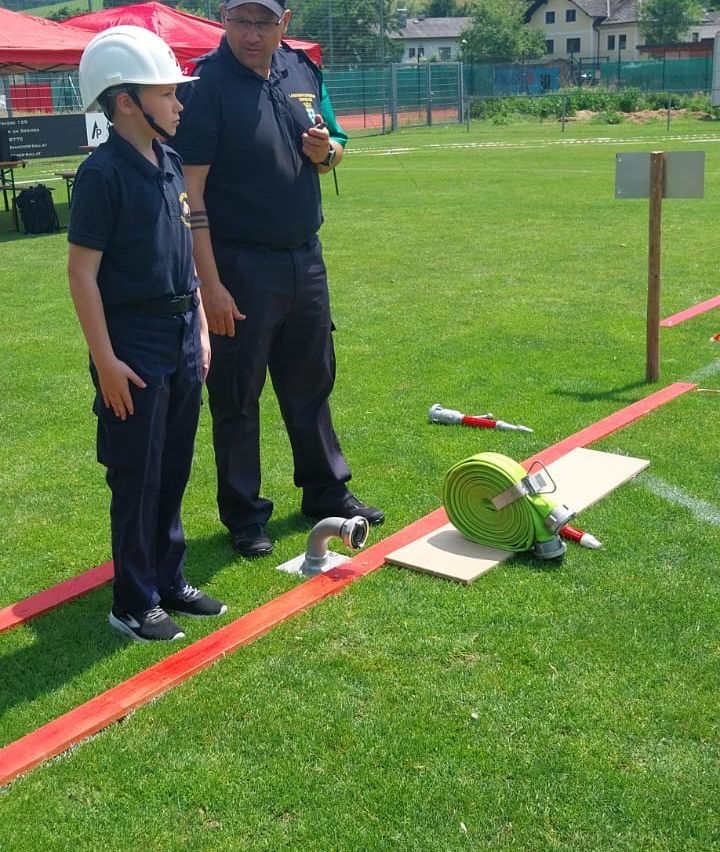 Ein Junge in einem Schutzhelm steht auf einem Feld, ein Feuerwehrmann steht hinter ihm. In der Nähe liegen ein Feuerwehrschlauch und ein Schlauchanschluss auf einer Pappplatte.