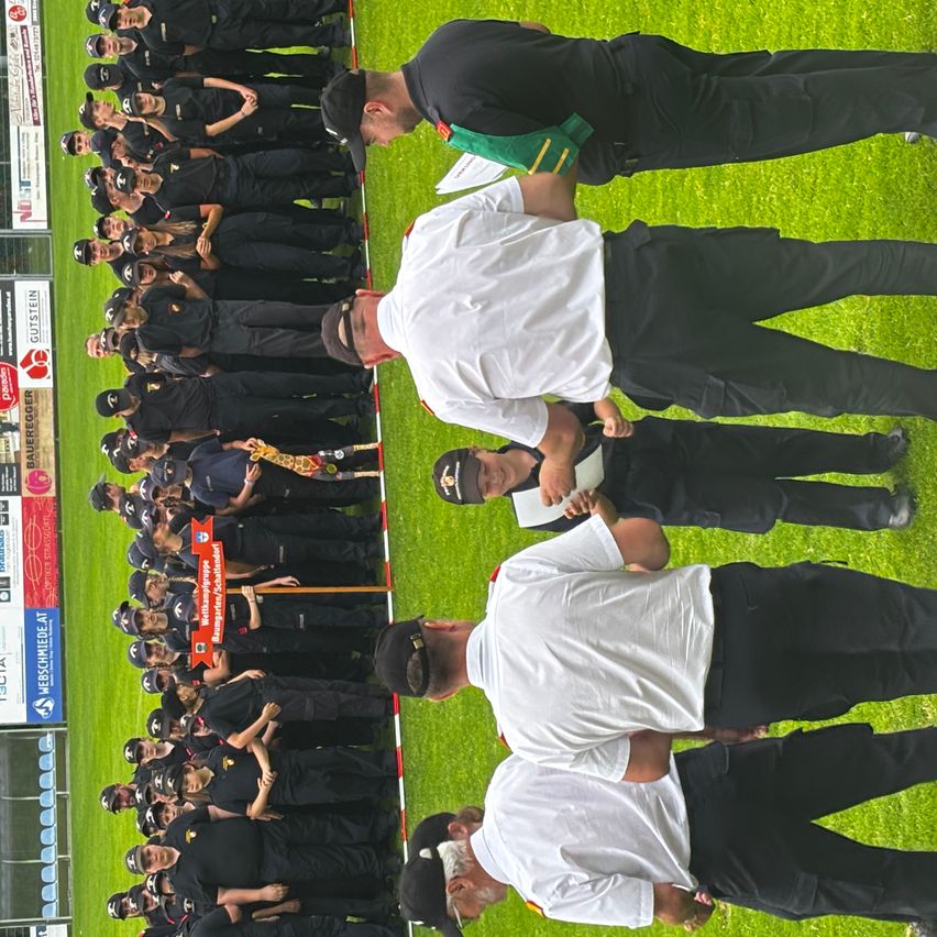 Eine Gruppe von Personen in Uniform steht in einer Reihe auf einem Rasenfeld, möglicherweise bei einer Veranstaltung. Sie werden von mehreren Personen in weißen Hemden und schwarzen Hosen begleitet. Einer der Personen hält ein Papier, während eine andere einen Stock hält. An den Seiten des Feldes befinden sich auch einige Werbungen.