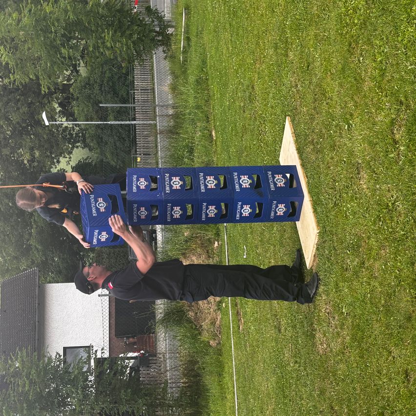 Zwei Personen auf einem Rasenfeld, eine lehnt sich an einen blauen Stapel Bierkisten, die andere schiebt ihn von hinten. Ein weißes Haus und eine Straßenlaterne sind im Hintergrund.