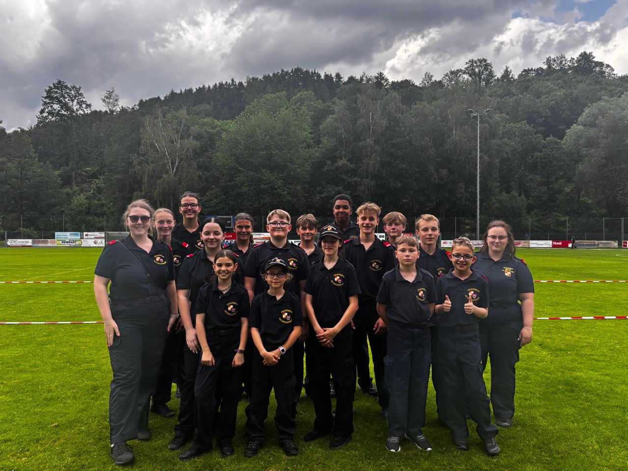 Eine Gruppe junger Personen in einheitlichen Outfits, einige mit Mützen, posiert für ein Foto auf einem Rasenfeld mit Waldhintergrund.