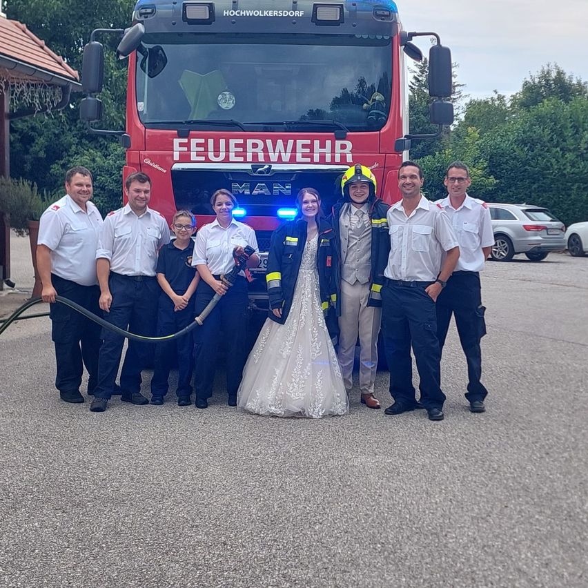 Eine Gruppe von Feuerwehrleuten und ein frisch verheiratetes Paar posiert vor einem roten Feuerwehrauto. Die Feuerwehrleute sind in Uniform und die Braut in einem Hochzeitskleid.