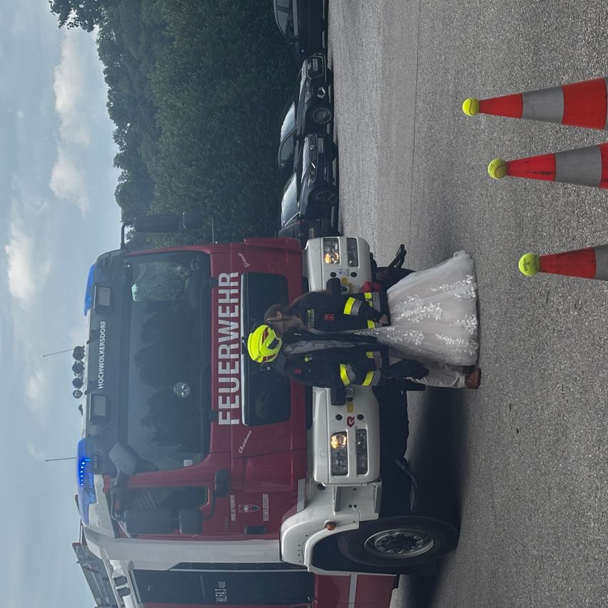 Ein Feuerwehrmann in Uniform steht neben einer Frau in einem Hochzeitskleid. Sie stehen vor einem roten Feuerwehrfahrzeug mit gelben und weißen Markierungen. Auf der Straße sind Verkehrskegel aufgestellt.