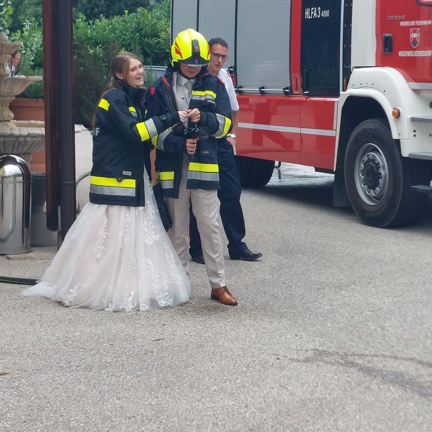 Eine Frau in einem Hochzeitskleid und zwei Feuerwehrleute stehen neben einem roten Feuerwehrauto. Die Feuerwehrleute halten Ferngläser und lächeln.