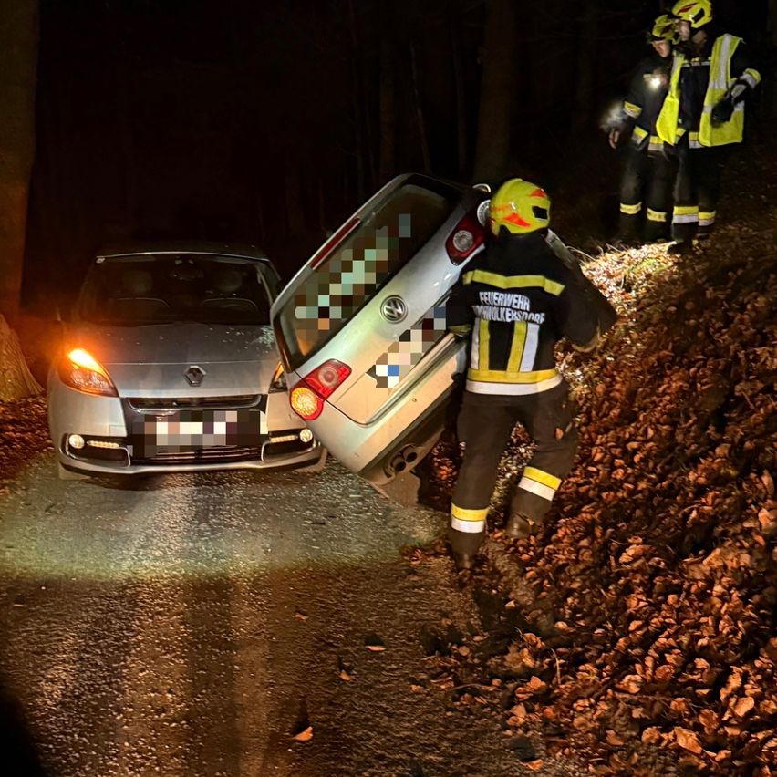 Ein Auto steht nachts am Straßenrand. Ein Feuerwehrmann hilft, das Auto anzuheben. Zwei weitere Feuerwehrleute stehen in der Nähe.