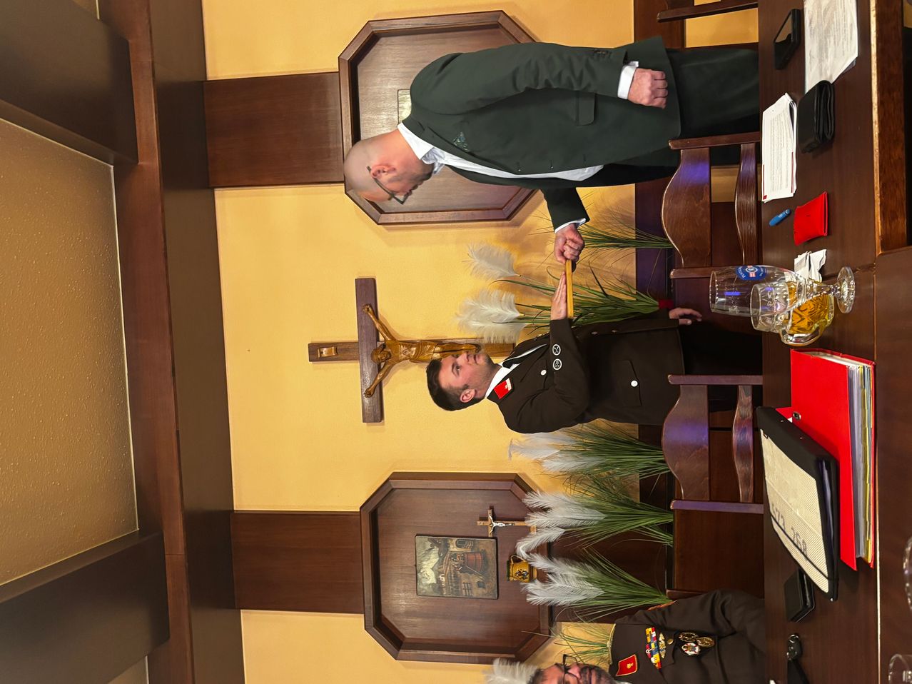 Ein Mann in Uniform erhält eine Auszeichnung von einem anderen Mann in einem Anzug, beide schauen nach unten. Die Szene ist drinnen, mit einem Kreuz an der Wand und einem Tisch mit Biergläsern.