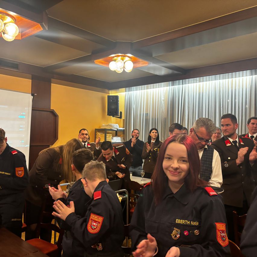 Eine Gruppe uniformierter Personen in einem Raum klatscht für eine lächelnde Frau in einer Uniform mit dem Namen Eberth Nadia. Der Raum hat gelbe Wände und Vorhänge.