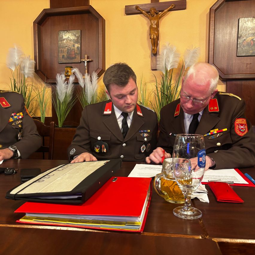 Drei Männer in Militäruniformen sitzen an einem Schreibtisch in einer Kirche. Papiere, ein Ordner und Gläser Bier stehen auf dem Tisch. Ein Kruzifix ist an der Wand hinter ihnen angebracht.
