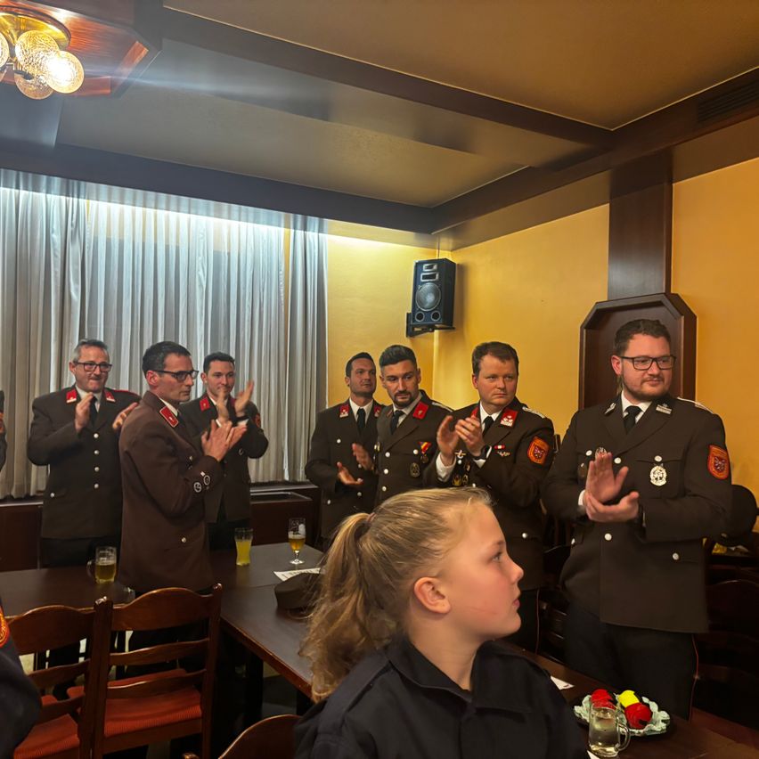 Mehrere uniformierte Personen klatschen in einem Raum mit gelben Wänden, Glastischen und Stühlen. Eine Frau sitzt ihnen gegenüber. An der Wand ist ein Lautsprecher angebracht.