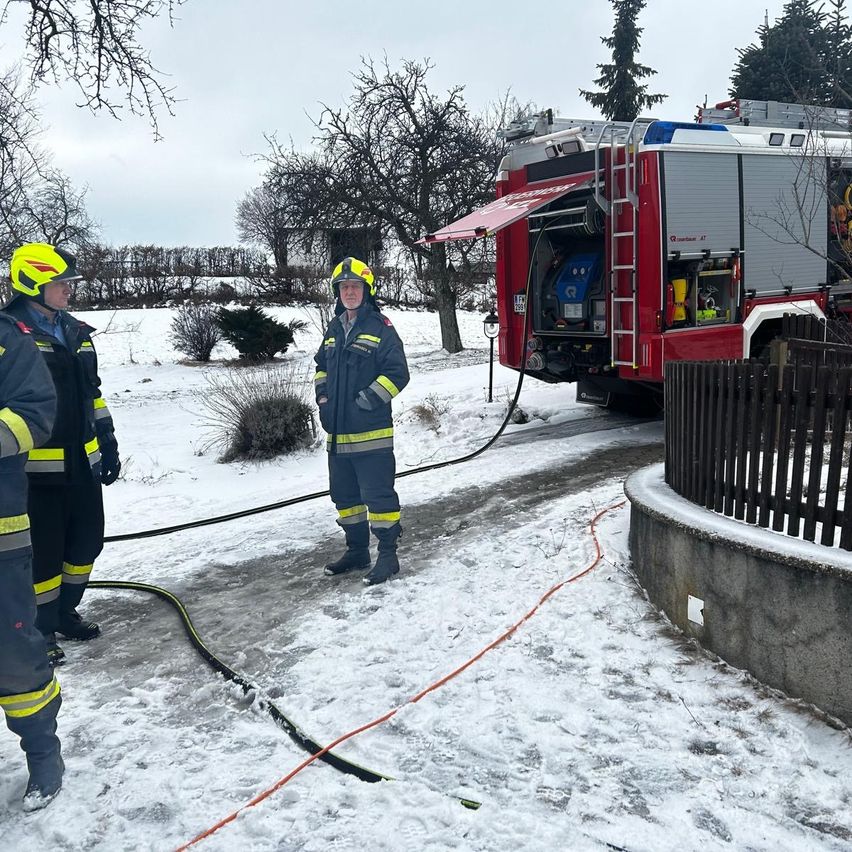 Drei Feuerwehrleute in Schutzausrüstung stehen im Schnee, in der Nähe eines roten Feuerwehrwagens mit offener Tür. Ein roter Schlauch liegt auf dem verschneiten Boden.