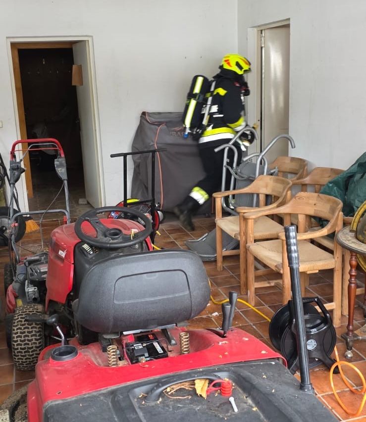 Ein Feuerwehrmann in voller Ausrüstung steht in einem Raum mit einem roten Rasenmäher, Stühlen und einem Tisch mit einer Schlauchrolle. Der Raum hat weiße Wände und geflieste Böden.