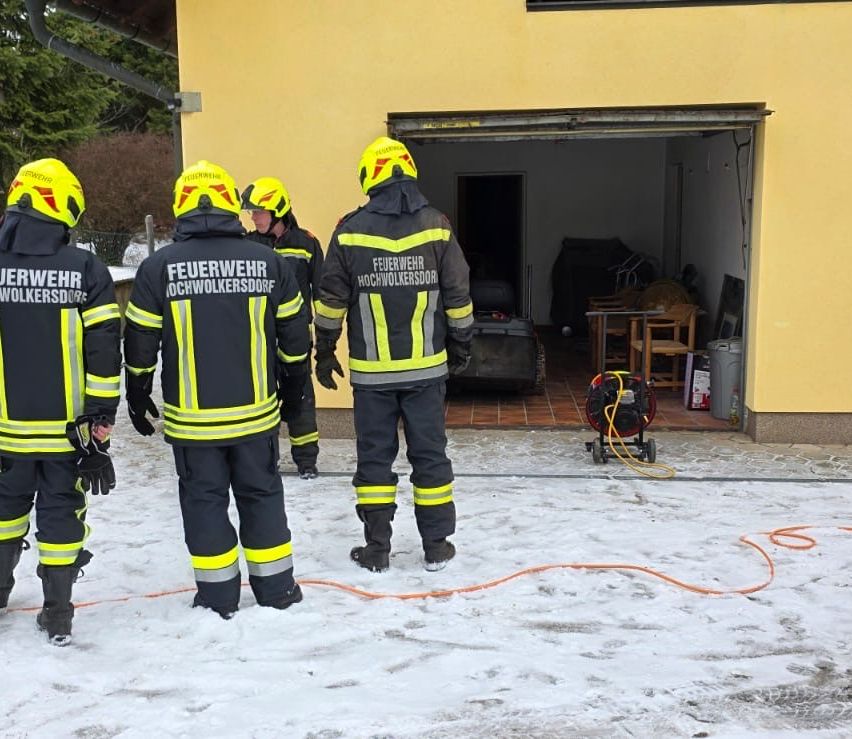 Vier Feuerwehrleute in Schutzkleidung stehen vor einem gelben Gebäude. Einer hält einen Schlauch, ein anderer einen Feuerlöscher. Die Szene ist verschneit.