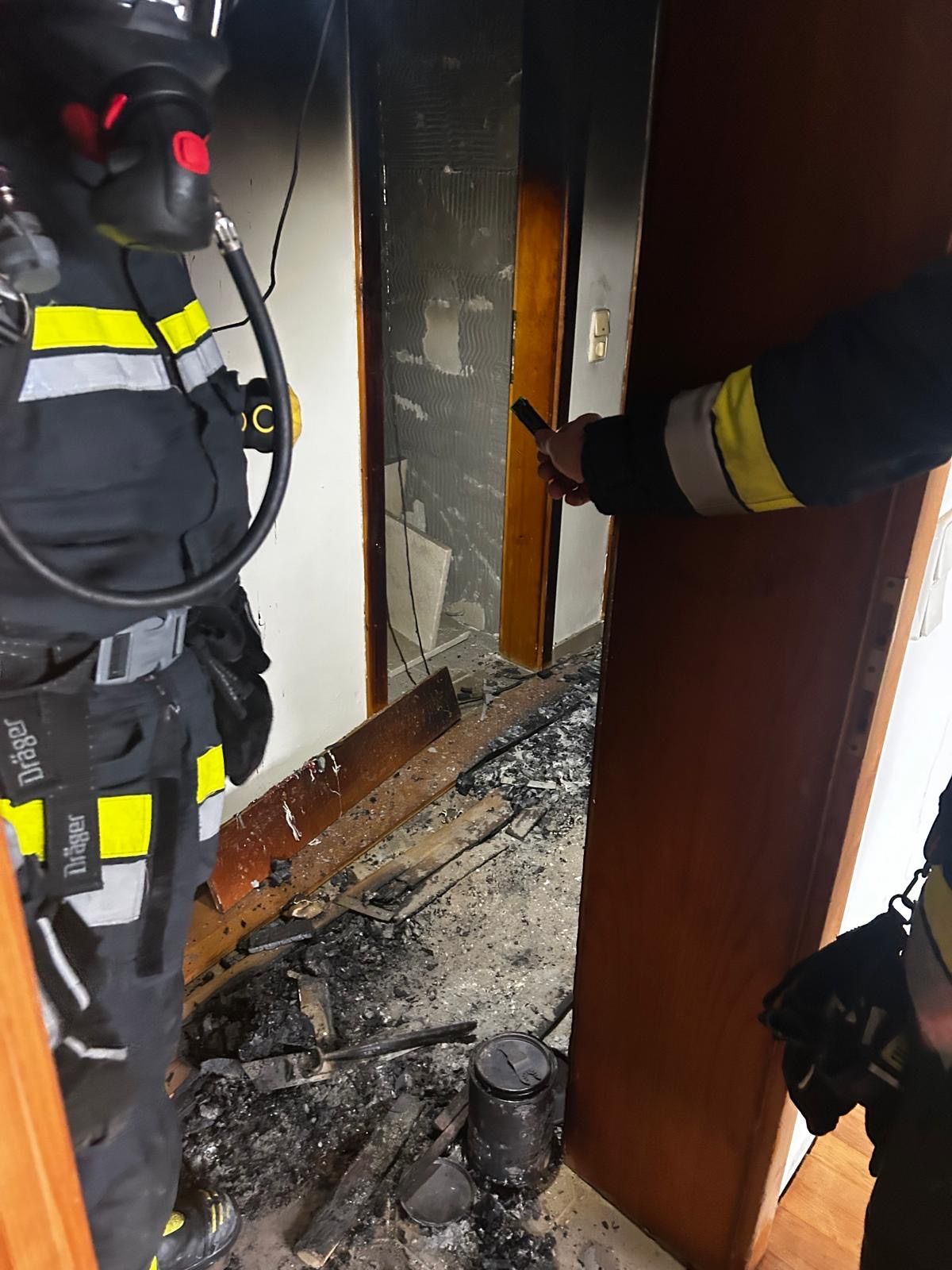 Feuerwehrleute inspizieren einen beschädigten Raum mit verbrannten Wänden und Schutt, einer hält eine Taschenlampe.