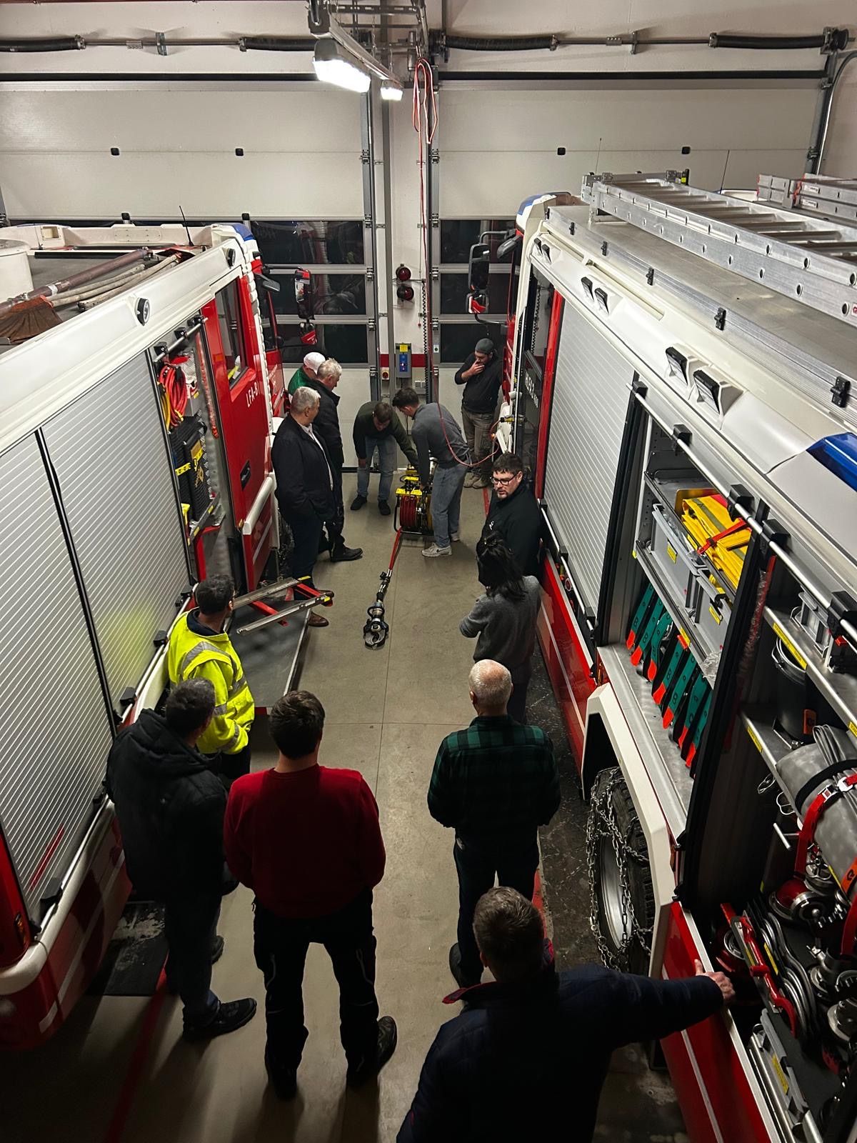 A group of people stand inside a fire station, inspecting fire trucks. One individual smokes a cigarette, while others examine equipment.