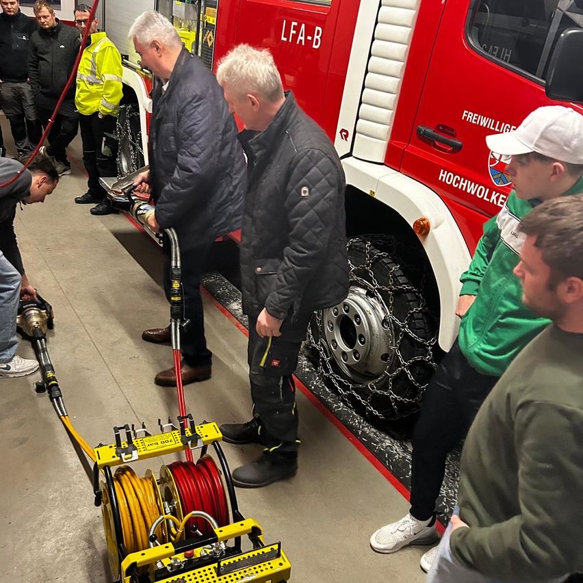 A group of people are gathered around a red fire truck. Two men are fixing something on the front of the truck, while others are watching. One man is holding a tool.