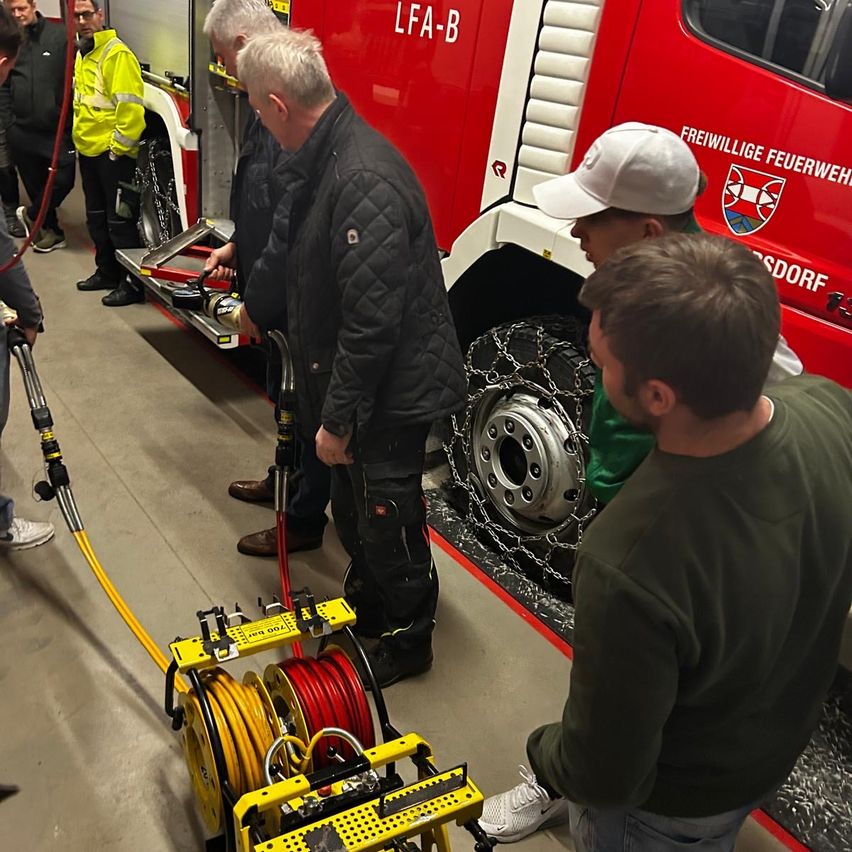 Several people gather around a red fire truck, focusing on a yellow machine and a wheel chain. A man in a white cap watches closely.