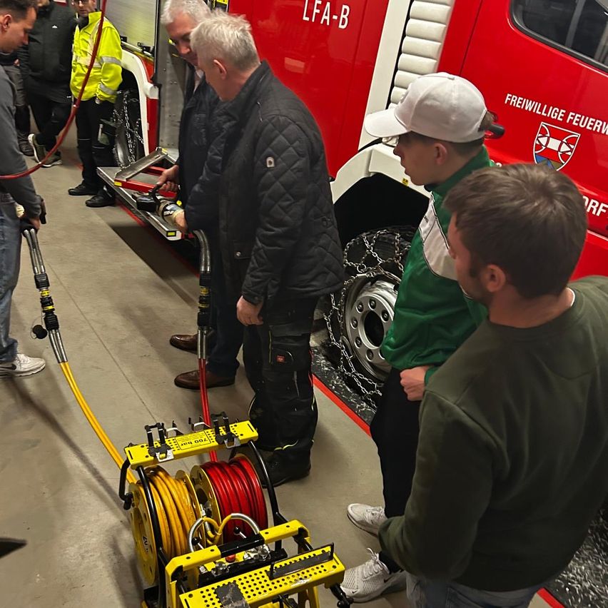 A group of men are gathered around a fire truck. One man in a green shirt is talking to another man in a white hat. There is a yellow machine with red and yellow hoses.