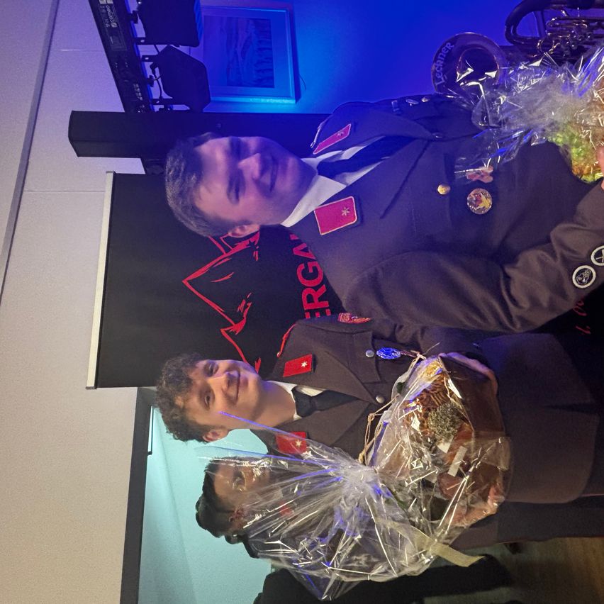 Two men in military uniforms are posing for a picture. One is holding a wrapped gift. Behind them is a wall with a black backdrop and red text.