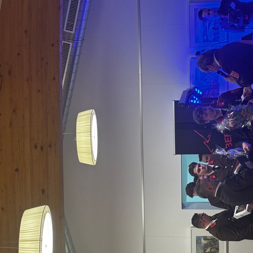 A group of men in uniforms gather around a stage, some holding awards, under spotlights. Two lamps are lit on the left.